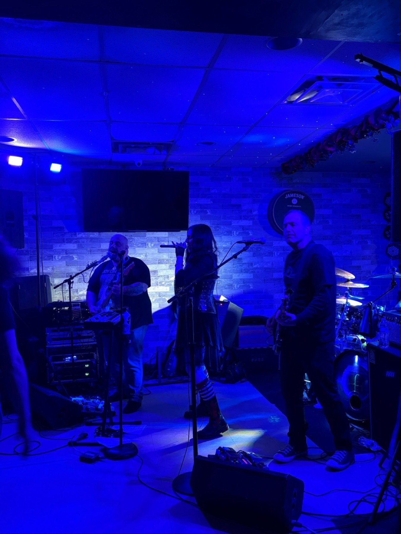 On a blue lit floor-level stage, a woman in a black lace peasant sleeve dress and corset sings between a t-shirt clad guitarist and base player.