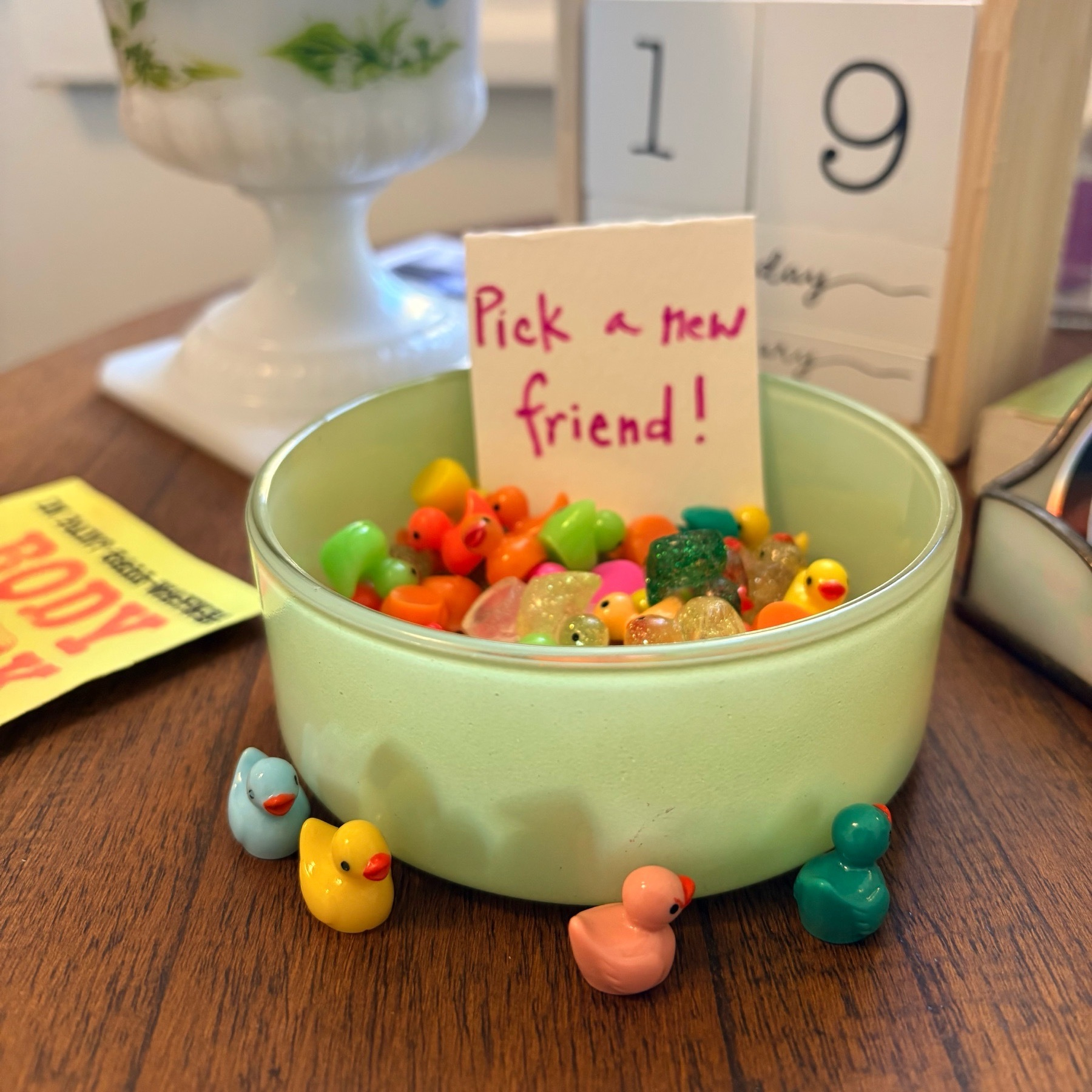 A green bowl filled with tiny plastic ducks in all colors sits on an end table, with a sign saying, “Pick a new friend!” There are other items in the table, and four tiny ducks along the bowl’s bottom edge.