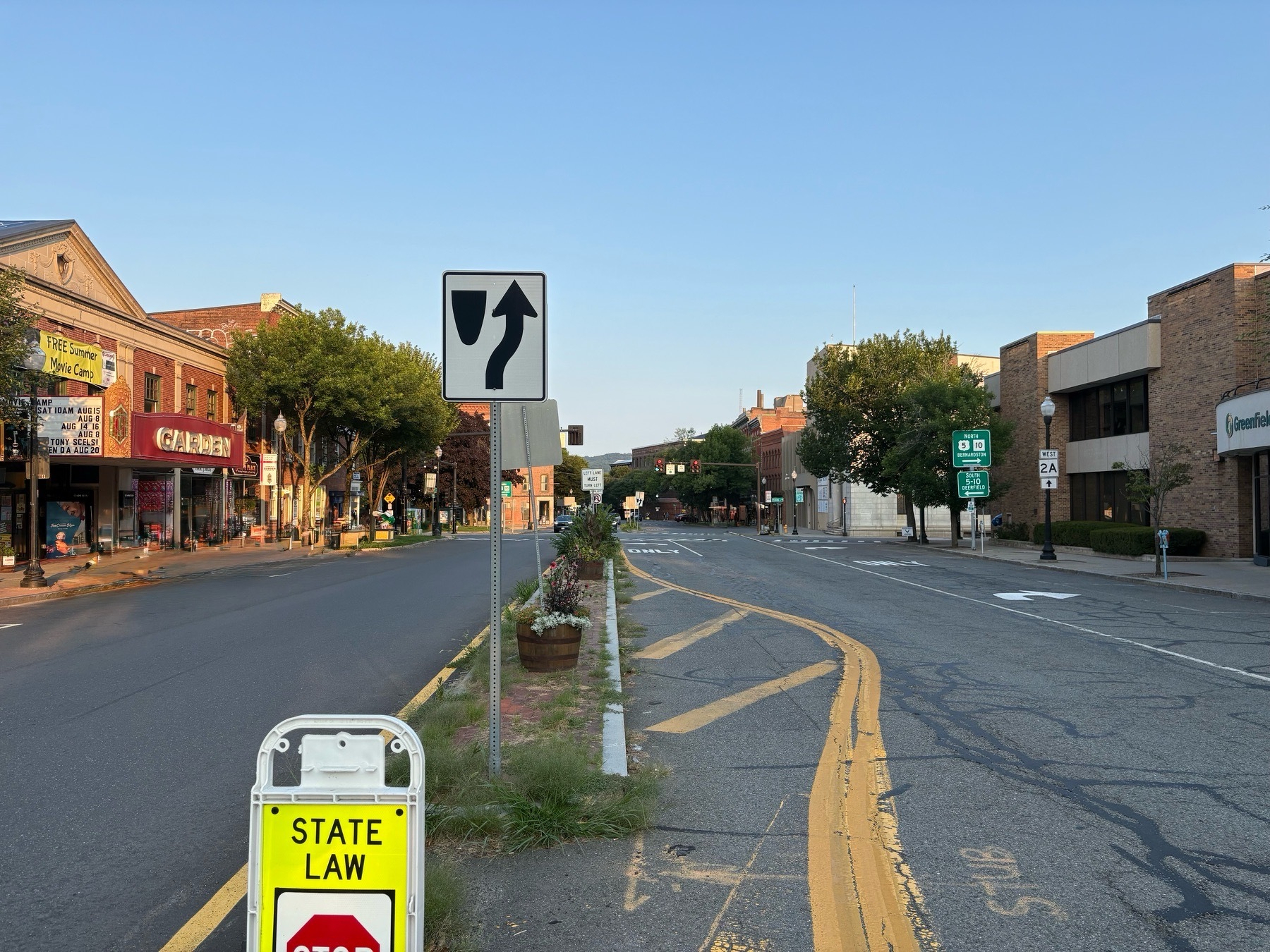 A quiet morning scene of a small town Main Street downtown, taken from the middle of the road. No cars are in the road. Morning sunlight touches the buildings on the left side, while the road and buildings on the right are in shadow. The morning sky is light blue.