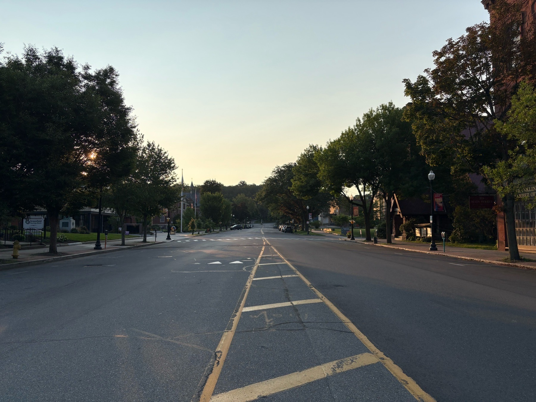 A Main Street in a small town’s downtown, with trees lining both sides of the road. The picture is taken in the middle of the road. Morning sunlight shines from the left, hitting trees and a brick building on the right, while all else is in shadow. The sky is light blue.