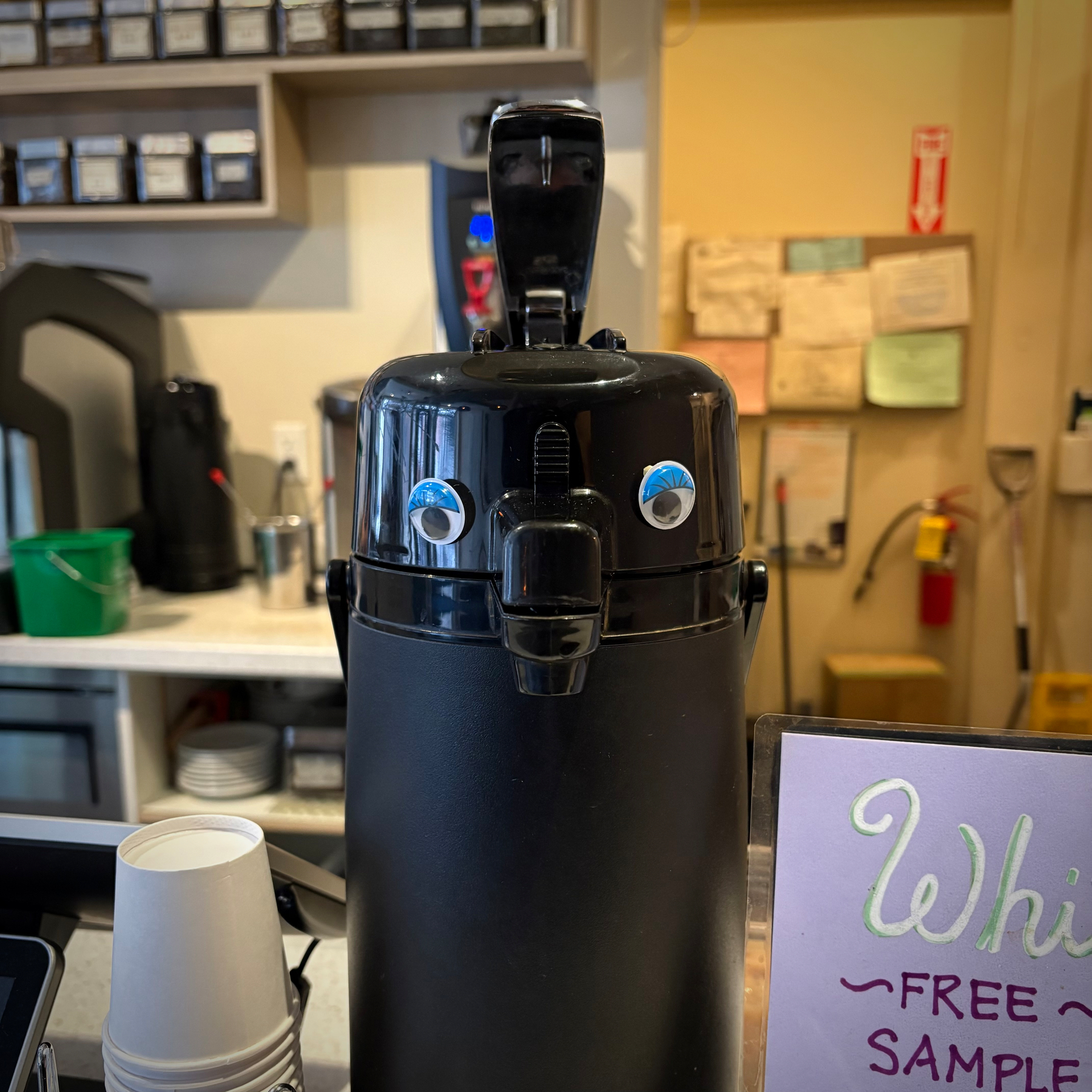 A black coffee dispenser is placed on a counter. A pair of half-lidded googly eyes with eyelashes and are affixed around the spout, giving the appearance of a slightly tired face.