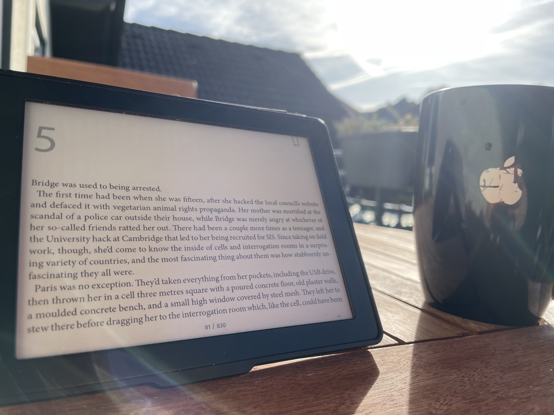 Tolino ebook reader in landscape, standing upright next to a coffee mug with a scratched up Apple logo. The ebook shows the beginning of Chapter 5. 