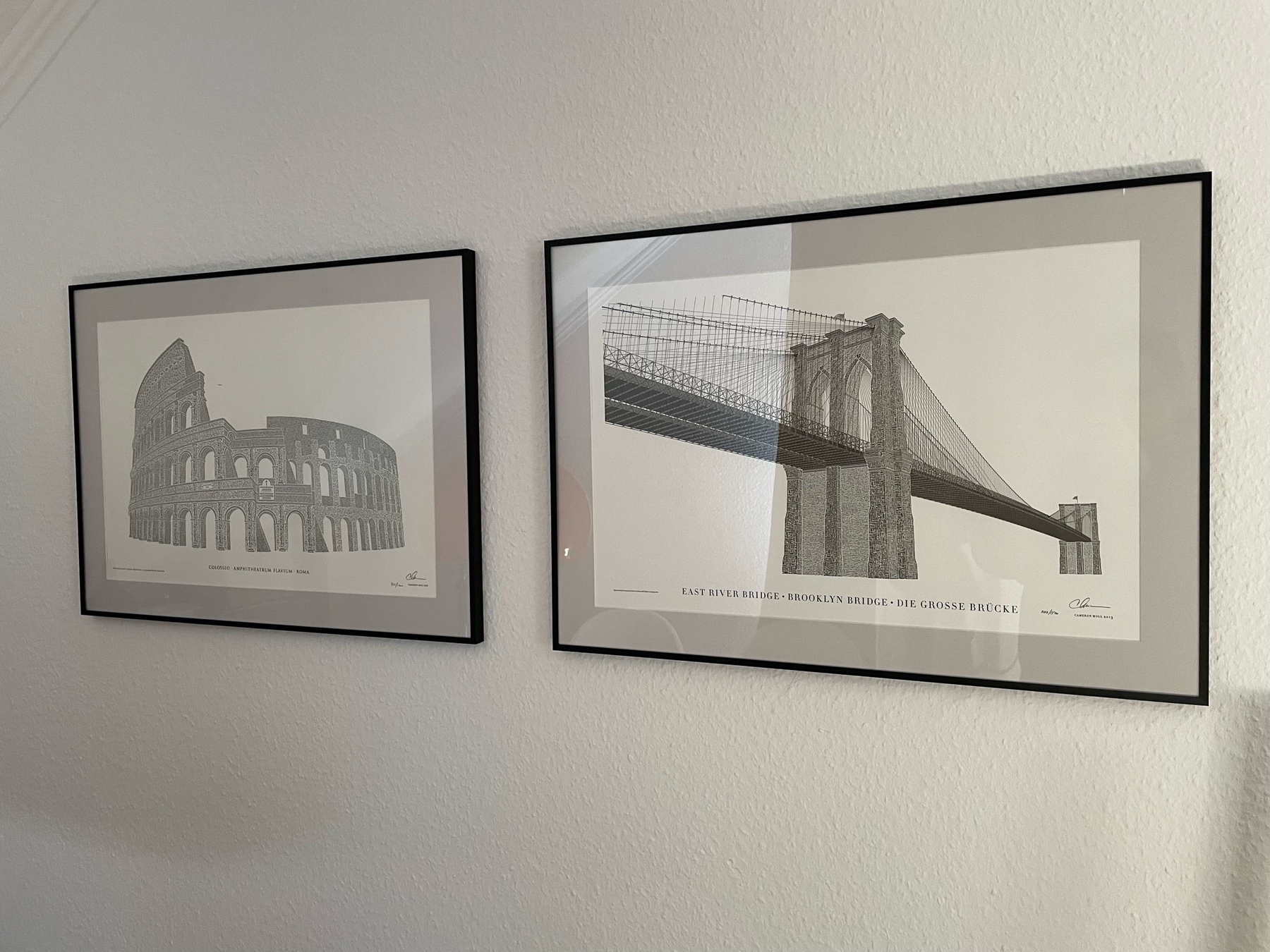 Photo of two prints hanging on a wall with a black frame around them. The left print displays the colloseum in Rome. The right one the brooklyn bridge. Both images are made out of letters. 