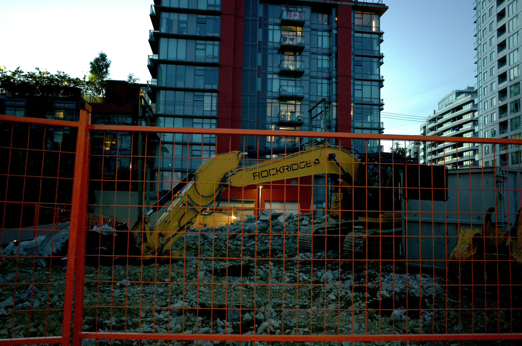 A yellow Rockridge excavator is parked behind an orange construction fence in front of a modern residential building.