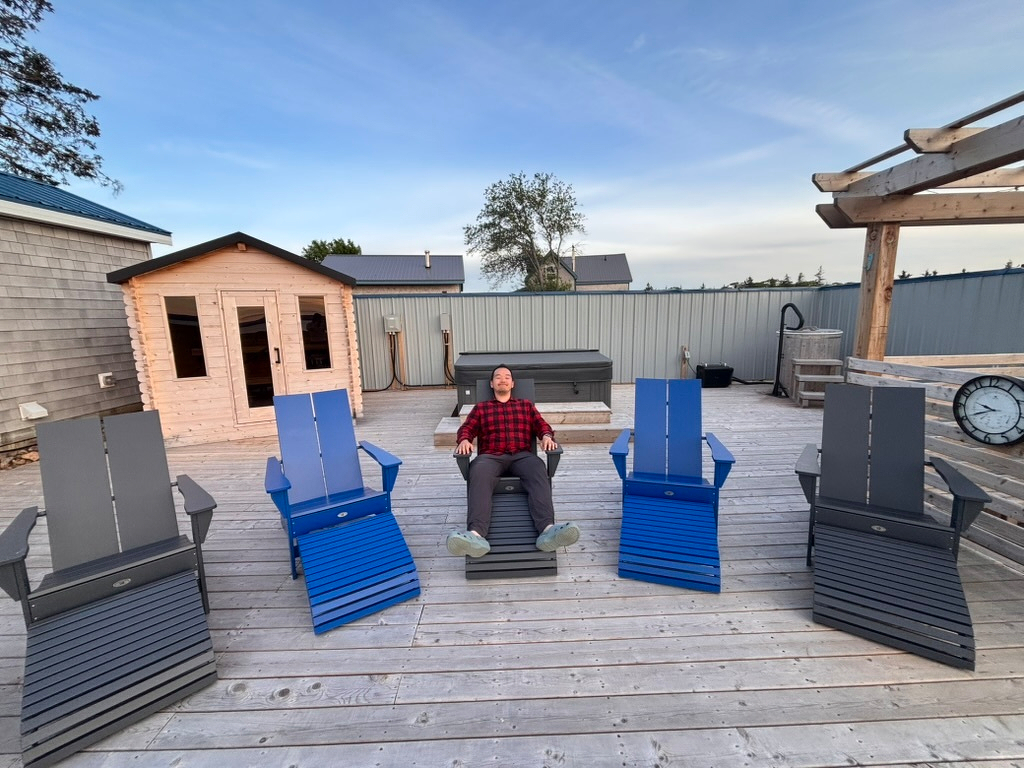 A person wearing a red and black plaid shirt is sitting and relaxing on a blue wooden chair on a spacious wooden deck surrounded by other blue and gray chairs, with a small shed and outdoor furniture in the background.