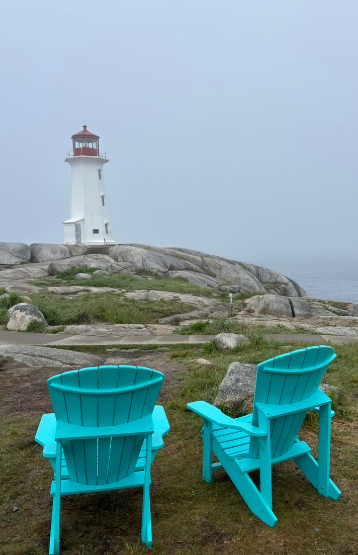 Two turquoise Adirondack chairs face a white lighthouse perched on rocky terrain under a foggy sky.