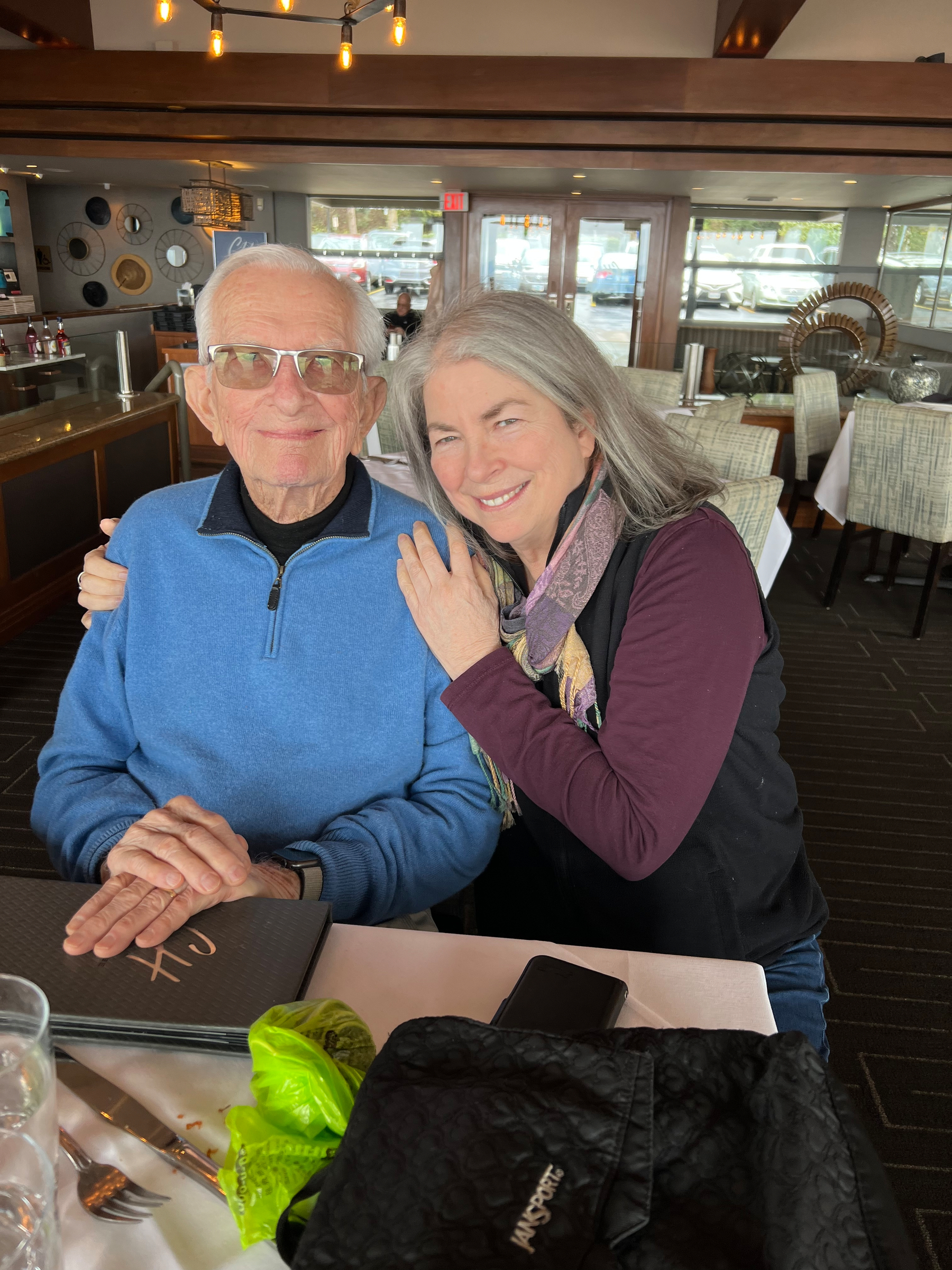 Elderly man in light blue sweater, hugged by a not-so-elderly woman with long gray hair in a purple sweater. 