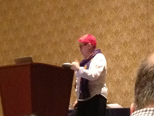 Auto-generated description: A person with red hair and wearing a scarf is standing at a podium in what appears to be a conference room.