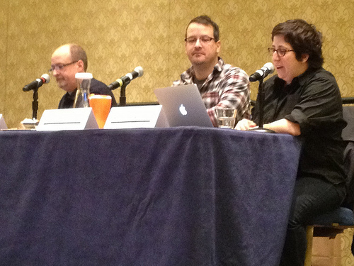 Auto-generated description: Three people are sitting at a table with microphones and laptops, possibly participating in a panel discussion or presentation.