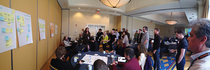 Auto-generated description: A group of people are gathered in a room for a meeting or brainstorming session, with notes displayed on the wall and several tables used for discussion.