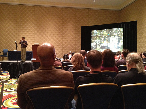 Auto-generated description: A speaker is presenting to an audience in a conference room, with a large screen displaying library shelves or bookcases.