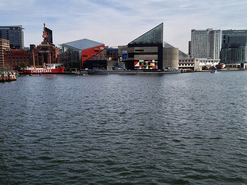 Auto-generated description: A waterfront scene features a mix of modern and historic buildings, a red ship named Chesapeake, and calm water under a partly cloudy sky.