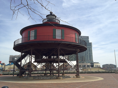 Auto-generated description: A round, red lighthouse sits elevated on struts with stairs leading up to it, set against an urban backdrop.