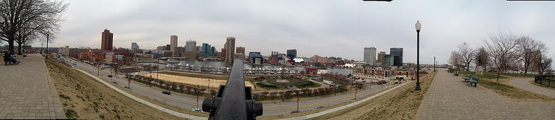 Auto-generated description: A panoramic view of a city skyline is captured from an elevated vantage point, showing buildings, waterfront area, and a park with benches and trees.