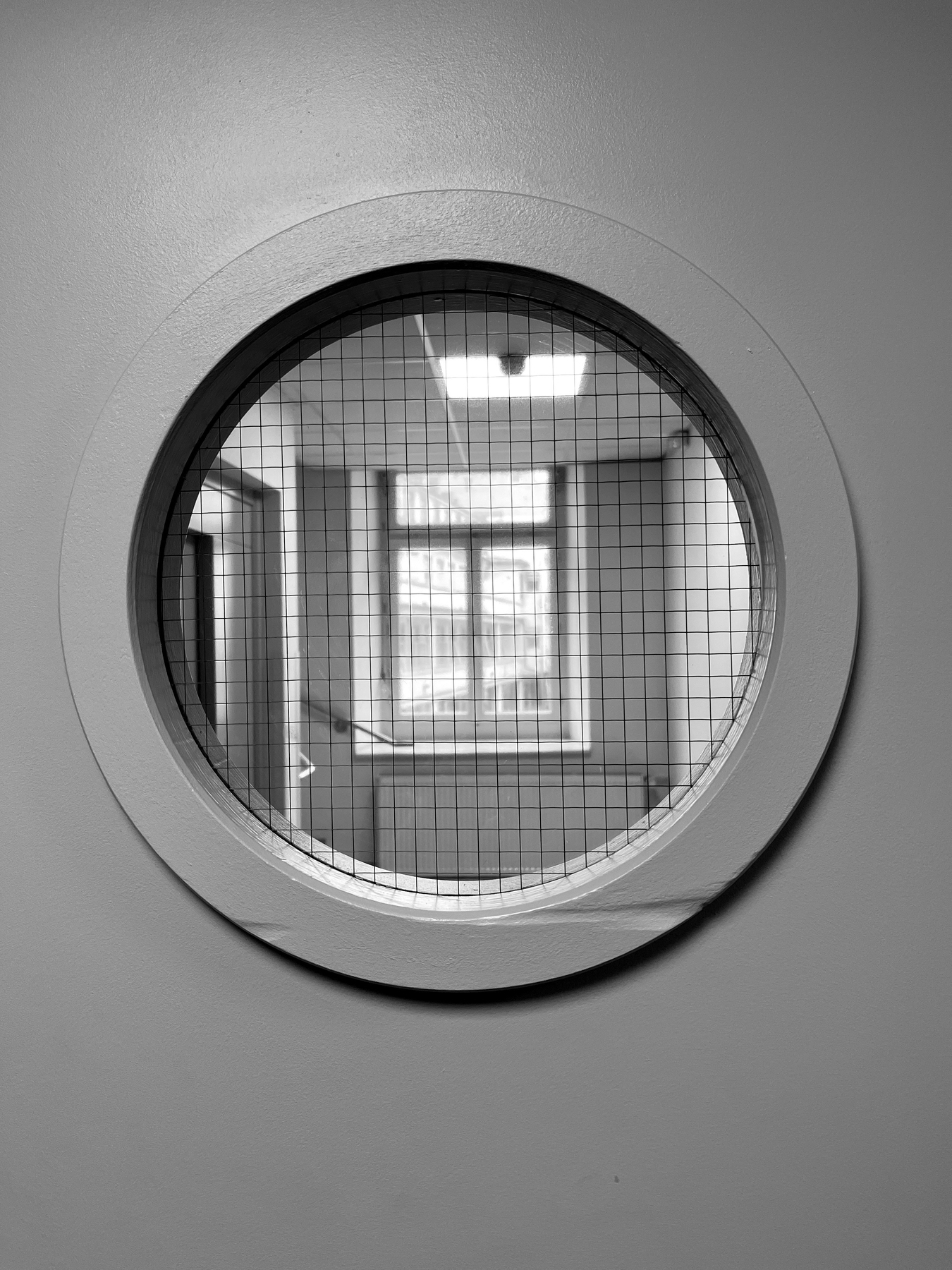 A black and white photo of a round window with a wire mesh, looking into another room with a window reflecting light.