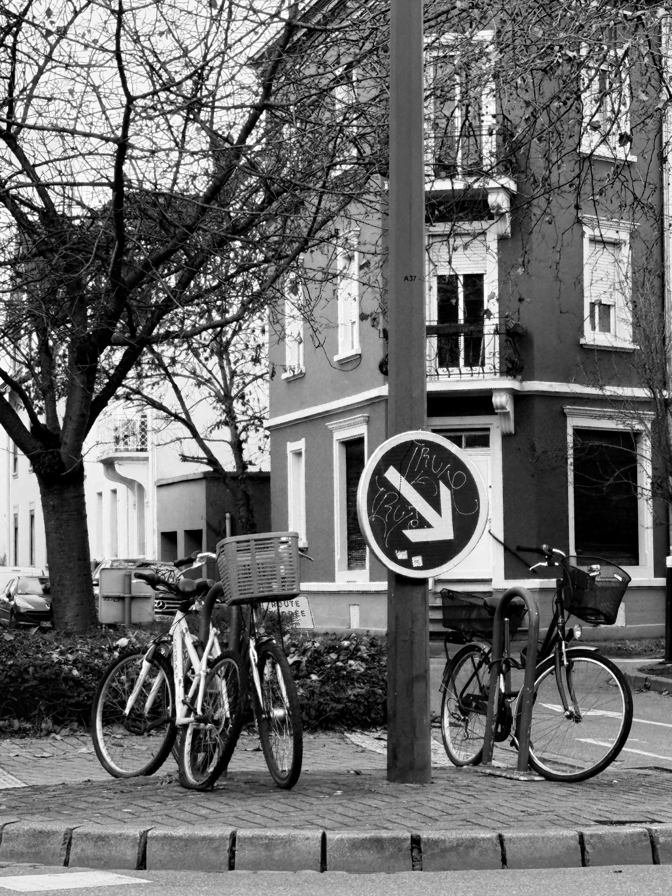 A black-and-white image featuring a few bicycles parked near a street sign indicating a one-way direction. In the background, a residential building with large windows and a tree bare of leaves is visible.