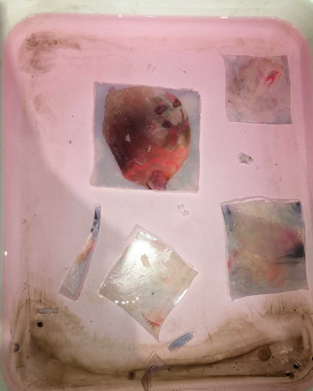 A pink tray contains several semi-transparent, irregularly shaped objects with splashes of color, set against a background with some smudges and stains.