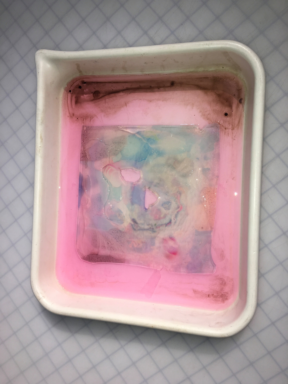 A square white dish with a pink liquid contains a partially submerged translucent material displaying a marbled pattern.
