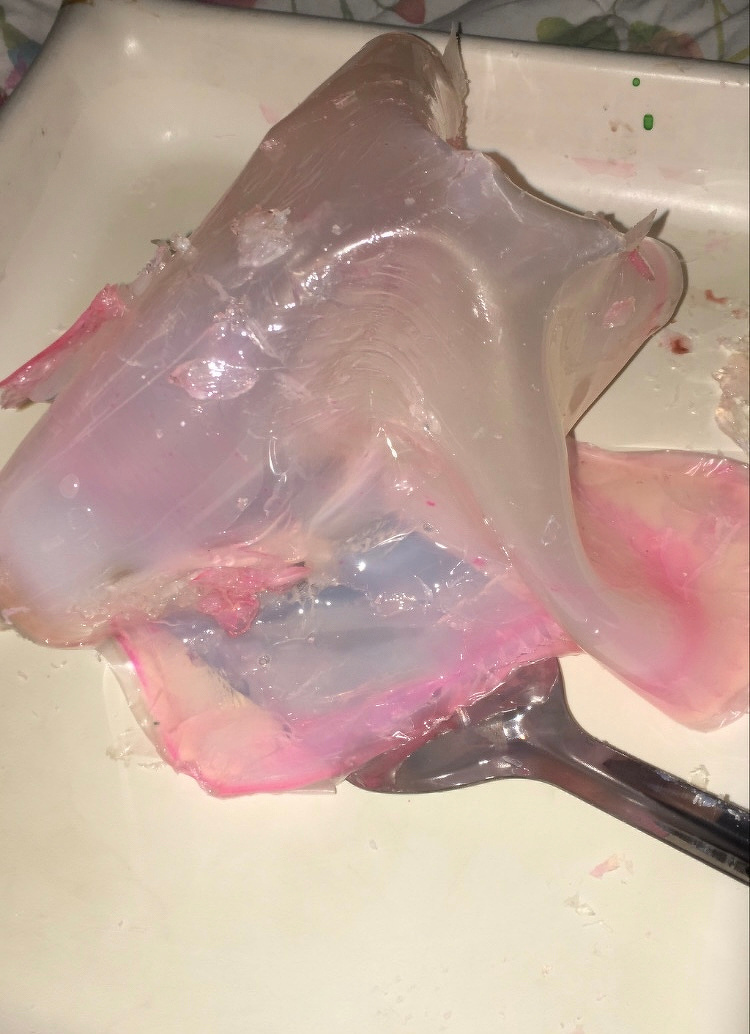 A translucent gelatinous substance with pink tinges is partially wrapped around a silver spoon on a beige surface.