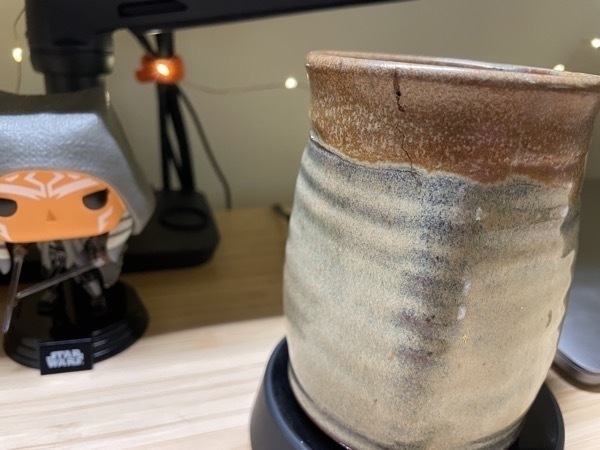 a clay mug with a crack in it, sitting on desk in front of a monitor and an Ahsoka bobblehead