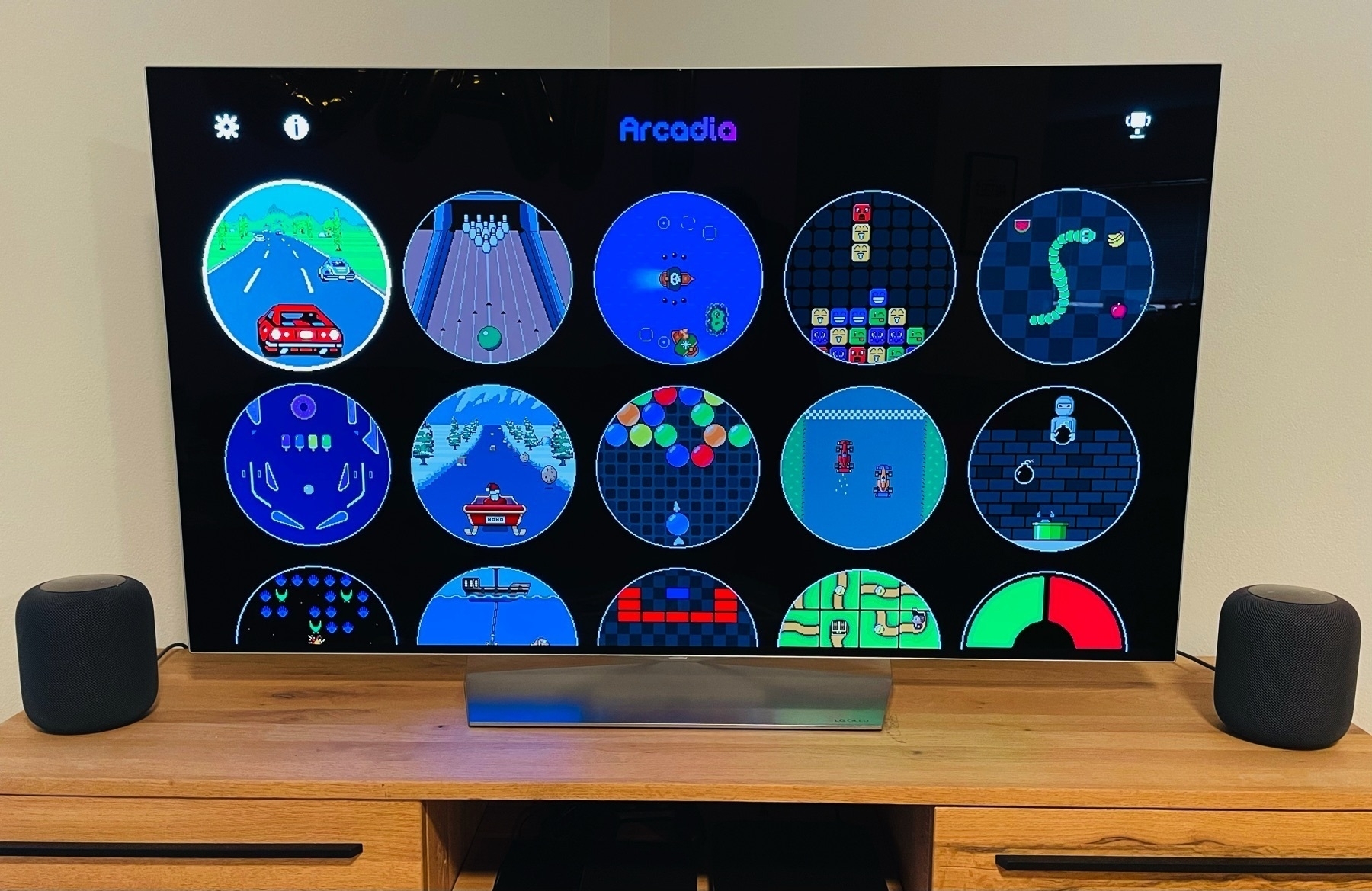 Arcadia June is shown running on an Apple TV 4K connected to an LG OLED TV, with the interface of games in a grid with circular icons. Two space grey HomePods in a stereo pair are also shown either side of the TV.