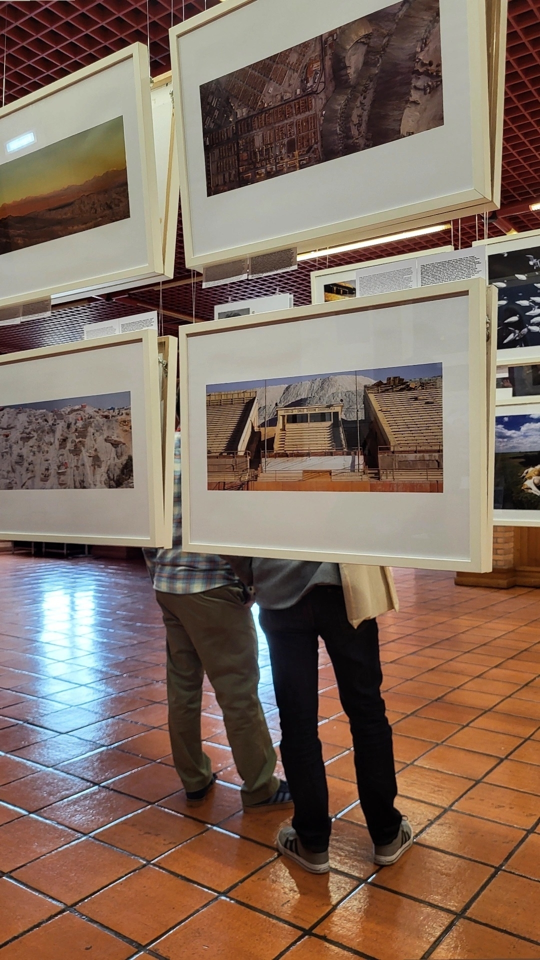 Two people are standing under a display of framed photographs in an art gallery with a tiled floor.