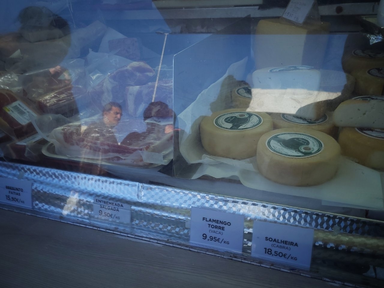 A display case shows various cheeses and meats with price tags, with people reflected on glass.