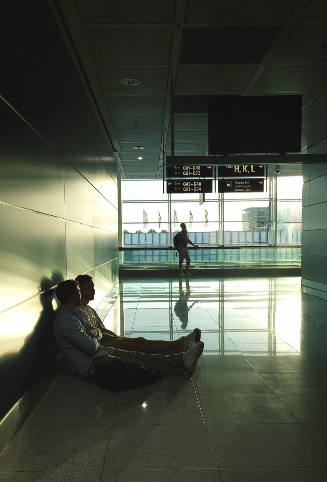 Men sitting at the floor at airport, with person walking in the back. 