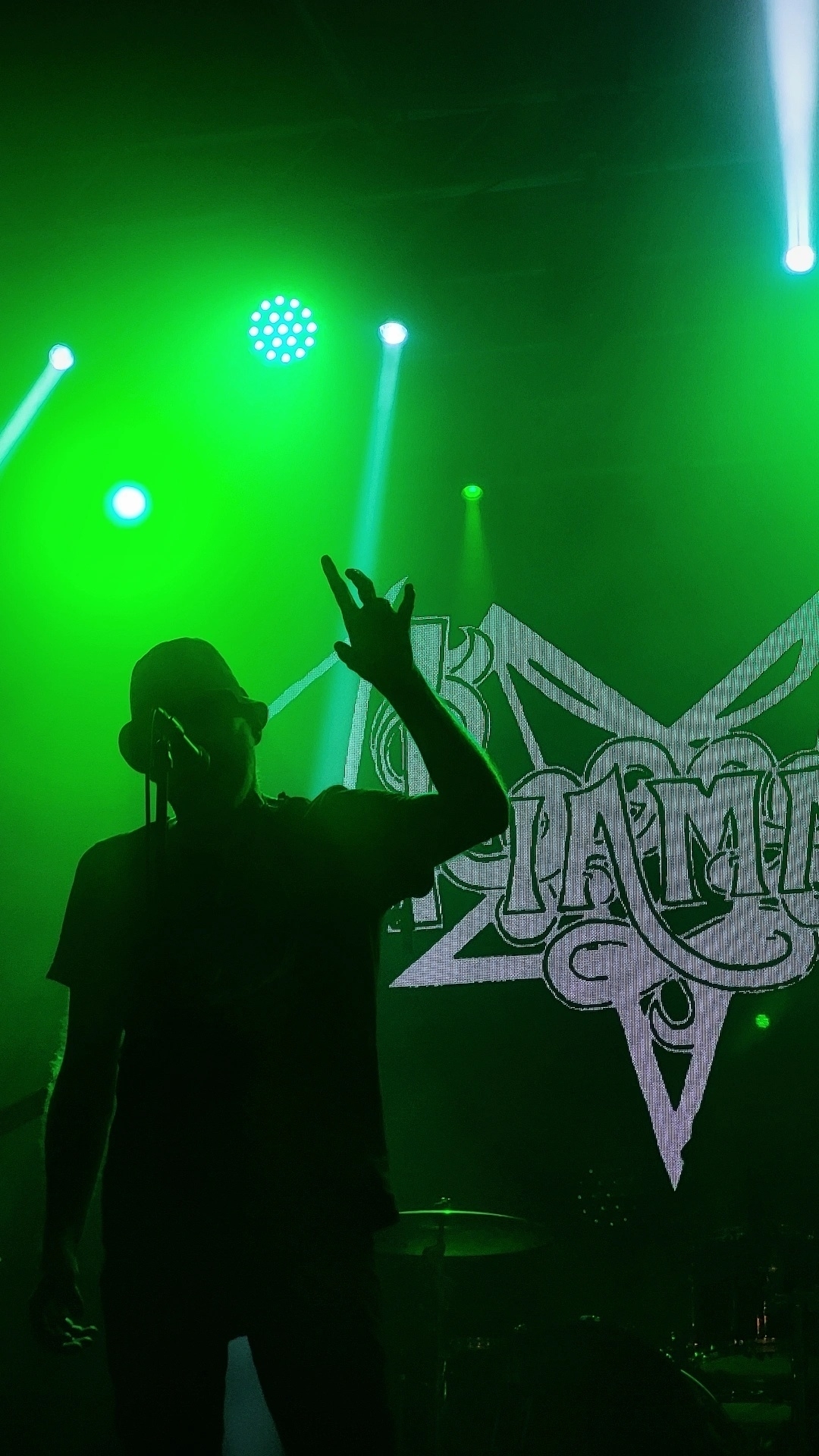 A silhouette of a performer stands on stage in front of a bright logo projected in the background, creating a dramatic atmosphere with dim lighting.