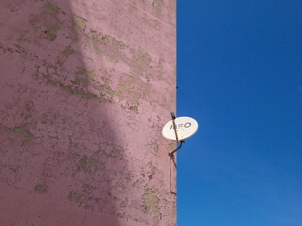 A satellite dish is mounted on a weathered pink wall against a clear blue sky.