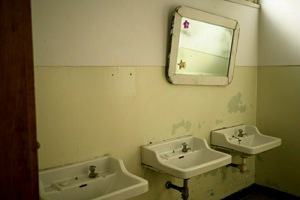 A dimly lit restroom features three white sinks aligned under a rectangular mirror with a small pink flower sticker.