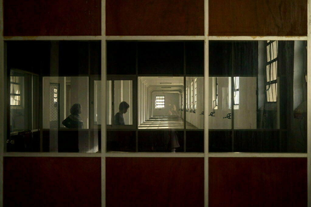 A silhouette of two people is seen through a corridor window with a row of windows on the right.