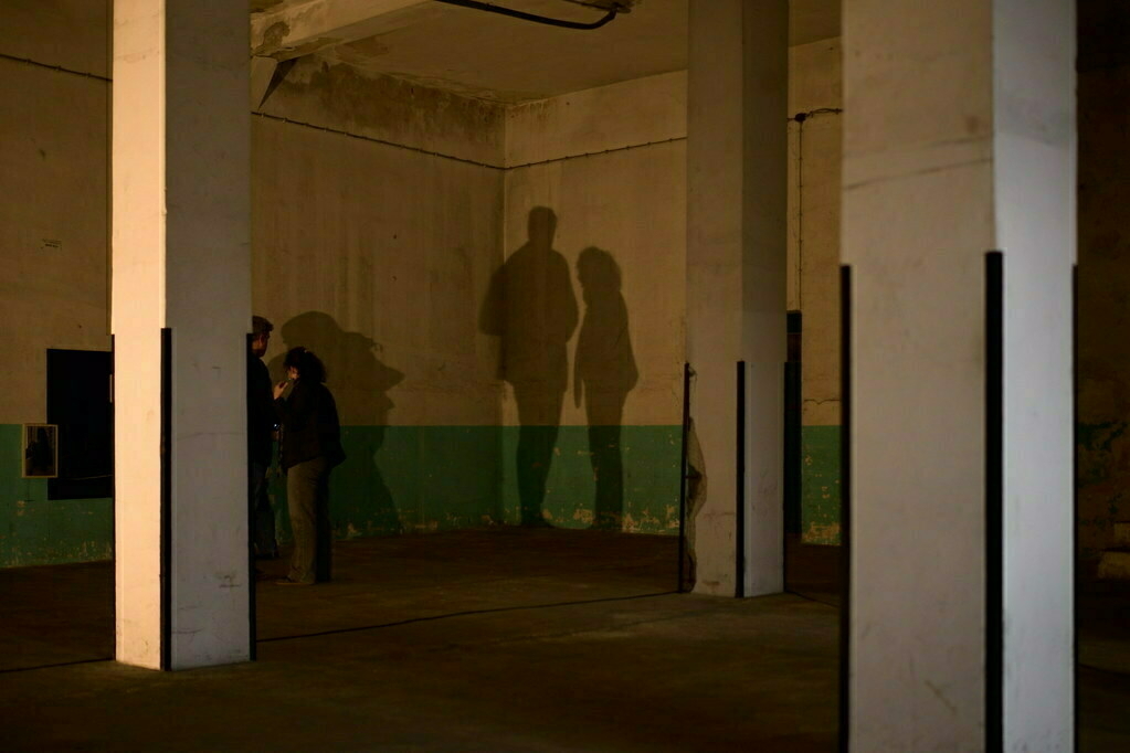 Two people stand in a dimly lit room with their shadows cast on a wall.