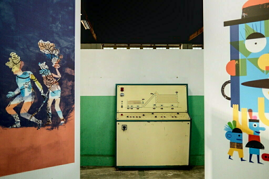 A beige control panel is set against a green and white wall between two colorful murals, one featuring two figures and the other abstract shapes and faces.