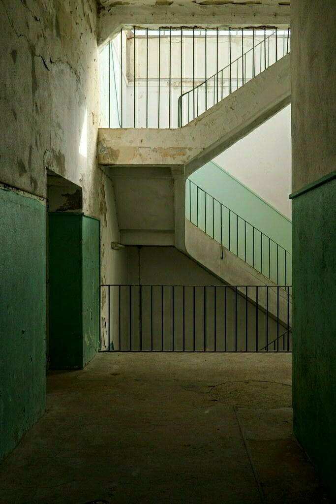 A dimly lit, empty stairwell with weathered green and white walls and a metal railing creates a sense of decay and abandonment.