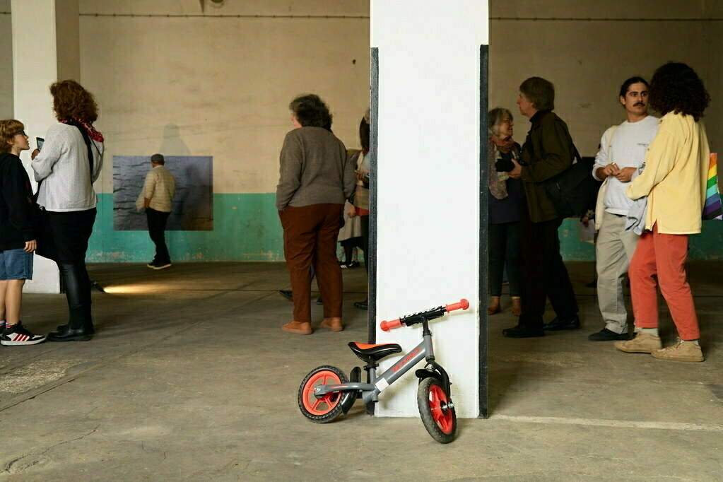 People are gathered in a room with artwork on the walls, and a small bicycle leans against a column.