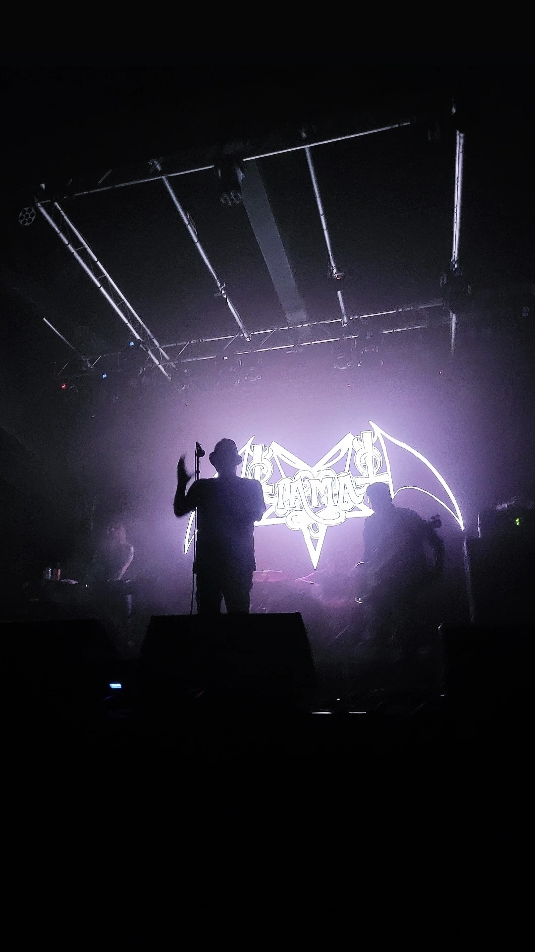 A silhouette of a performer stands on stage in front of a bright logo projected in the background, creating a dramatic atmosphere with dim lighting.