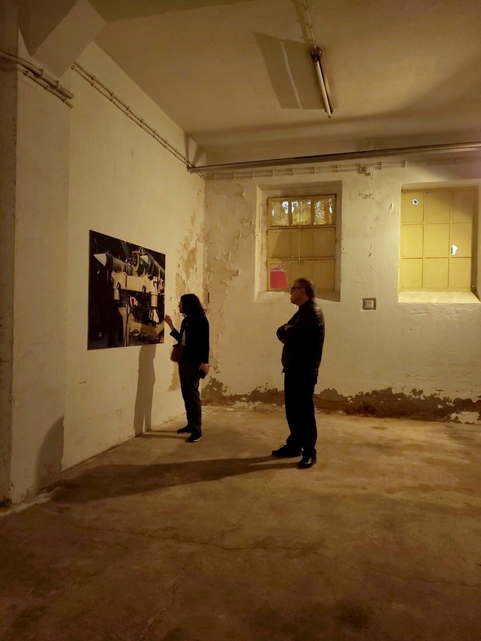 Two people are observing a piece of art in a dimly lit, industrial-style room with a worn appearance.