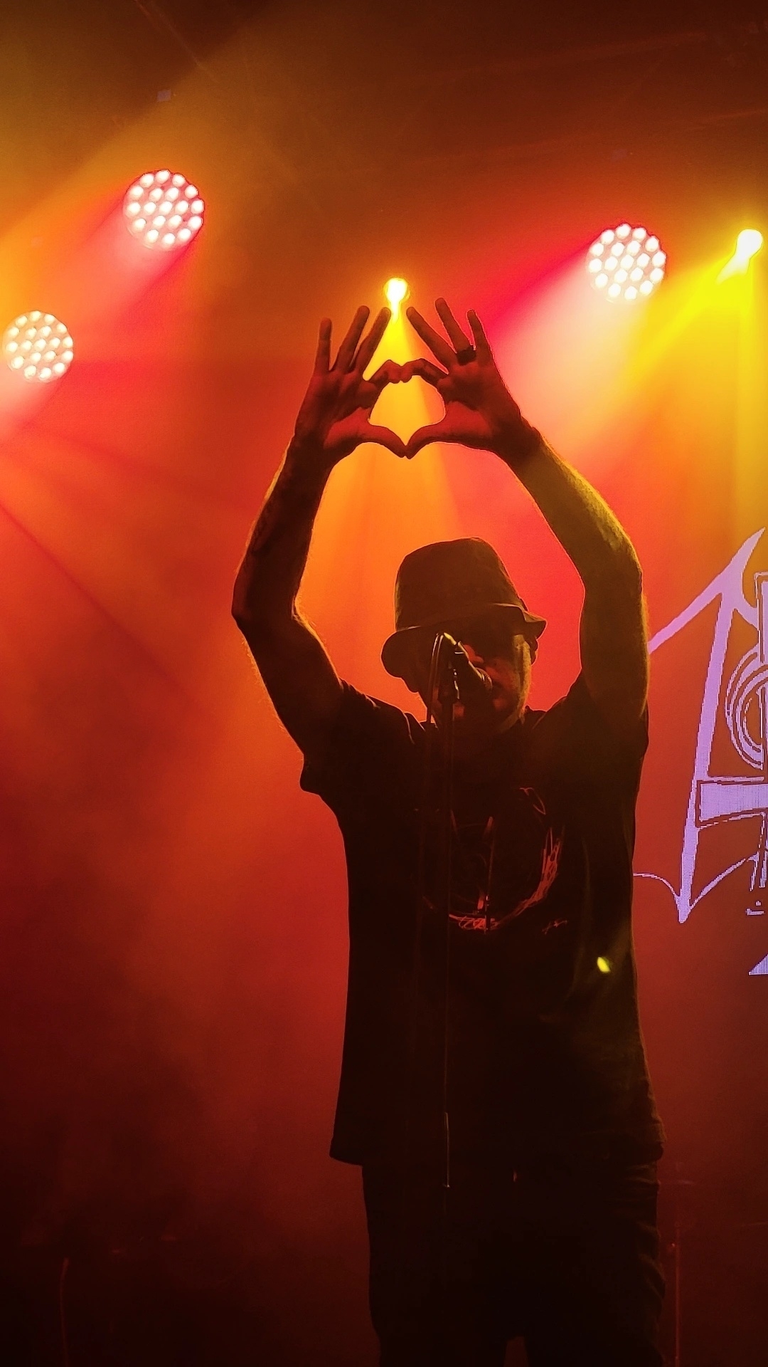 A performer on stage makes a heart shape with their hands under vibrant colored lights.