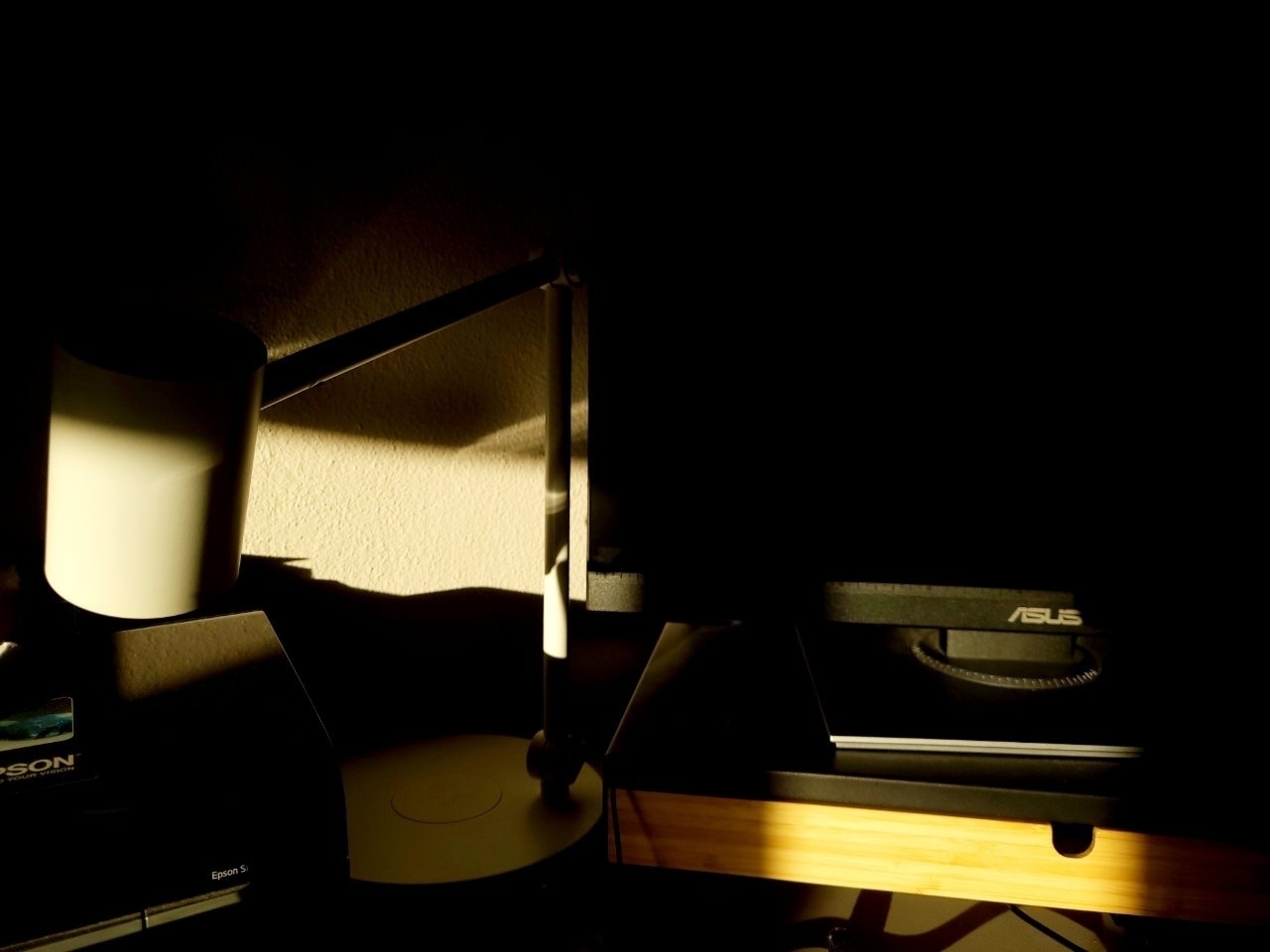 A dimly lit room features a desk lamp, a computer monitor, and a wooden table surface.