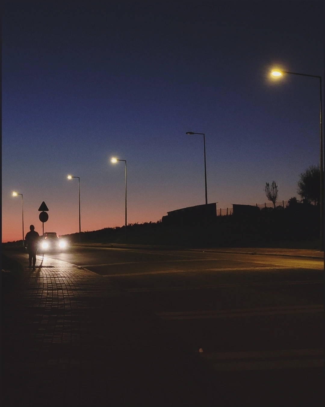 A quiet street scene at dusk features illuminated streetlights, a car with headlights on, and a silhouette of a person walking.