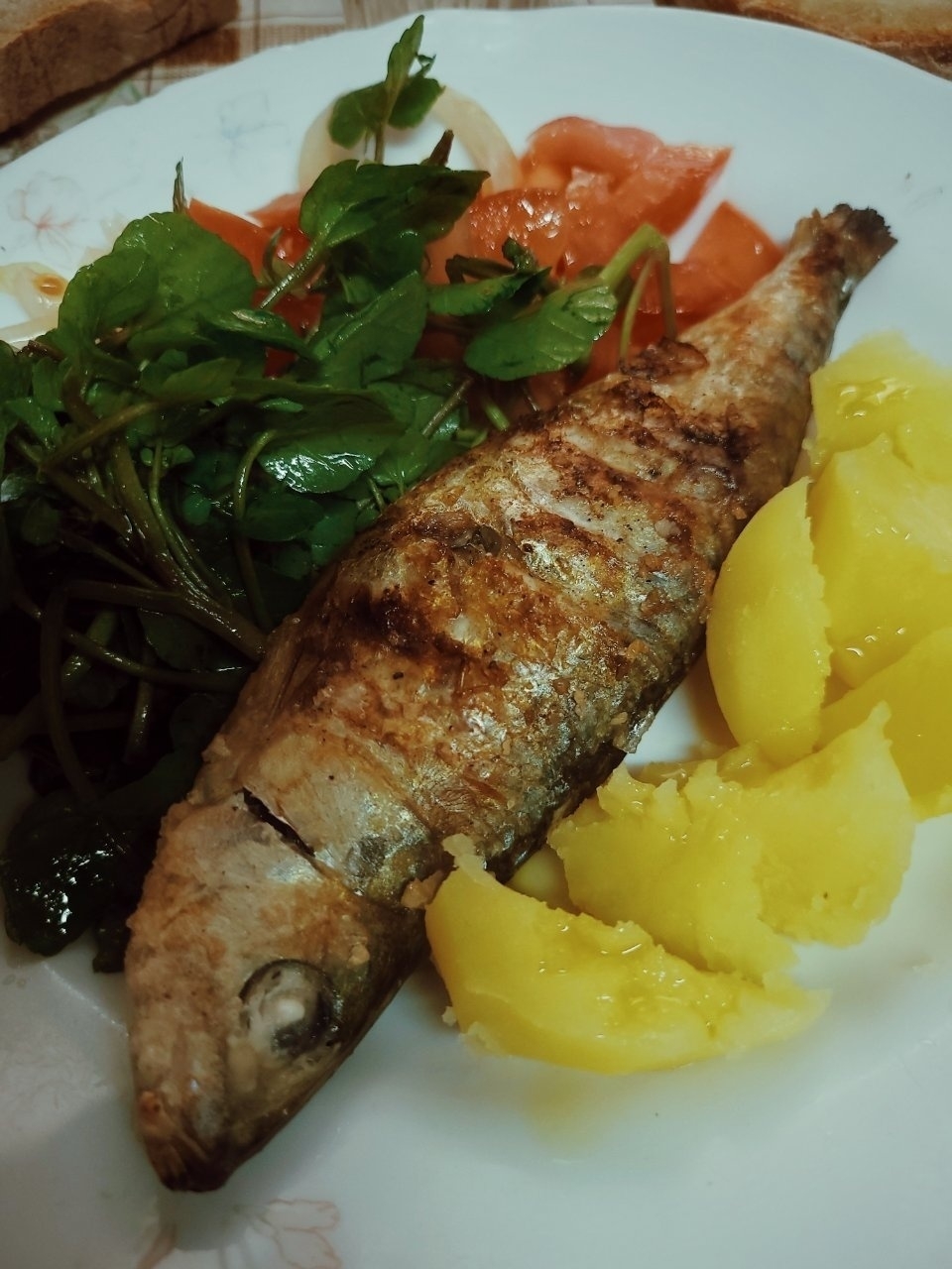 A grilled sardine is served with potatoes, leafy greens, and tomato slices on a white plate.