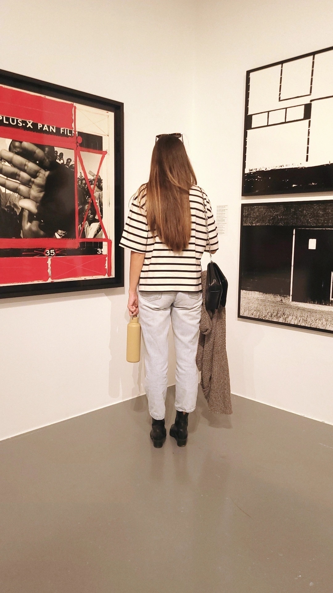 A woman with long hair, wearing a striped shirt and jeans, stands in a gallery observing abstract and black-and-white artwork.