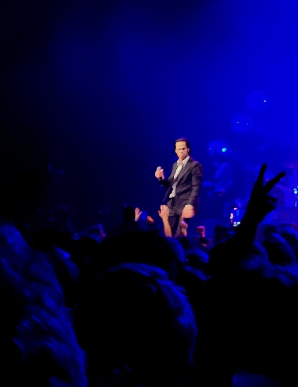 A performer onstage in a dark suit sings to an enthusiastic crowd under blue lighting.