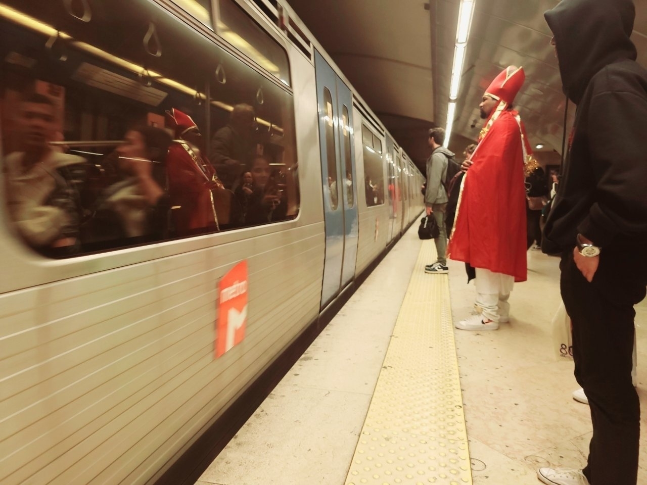 A person dressed in a bishop stands on a subway platform as a train arrives.