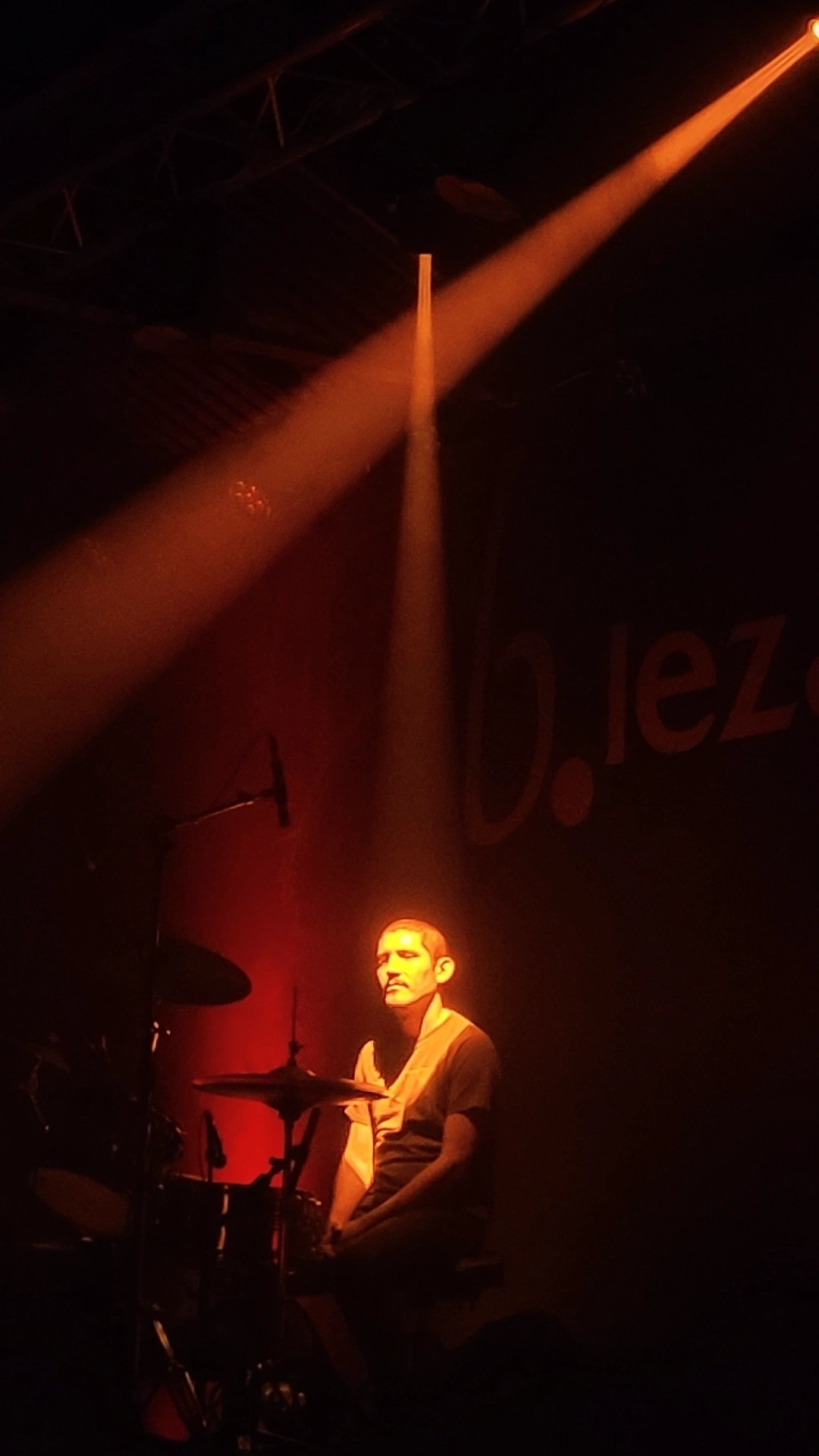 A drummer is illuminated by a warm spotlight on a dimly lit stage.