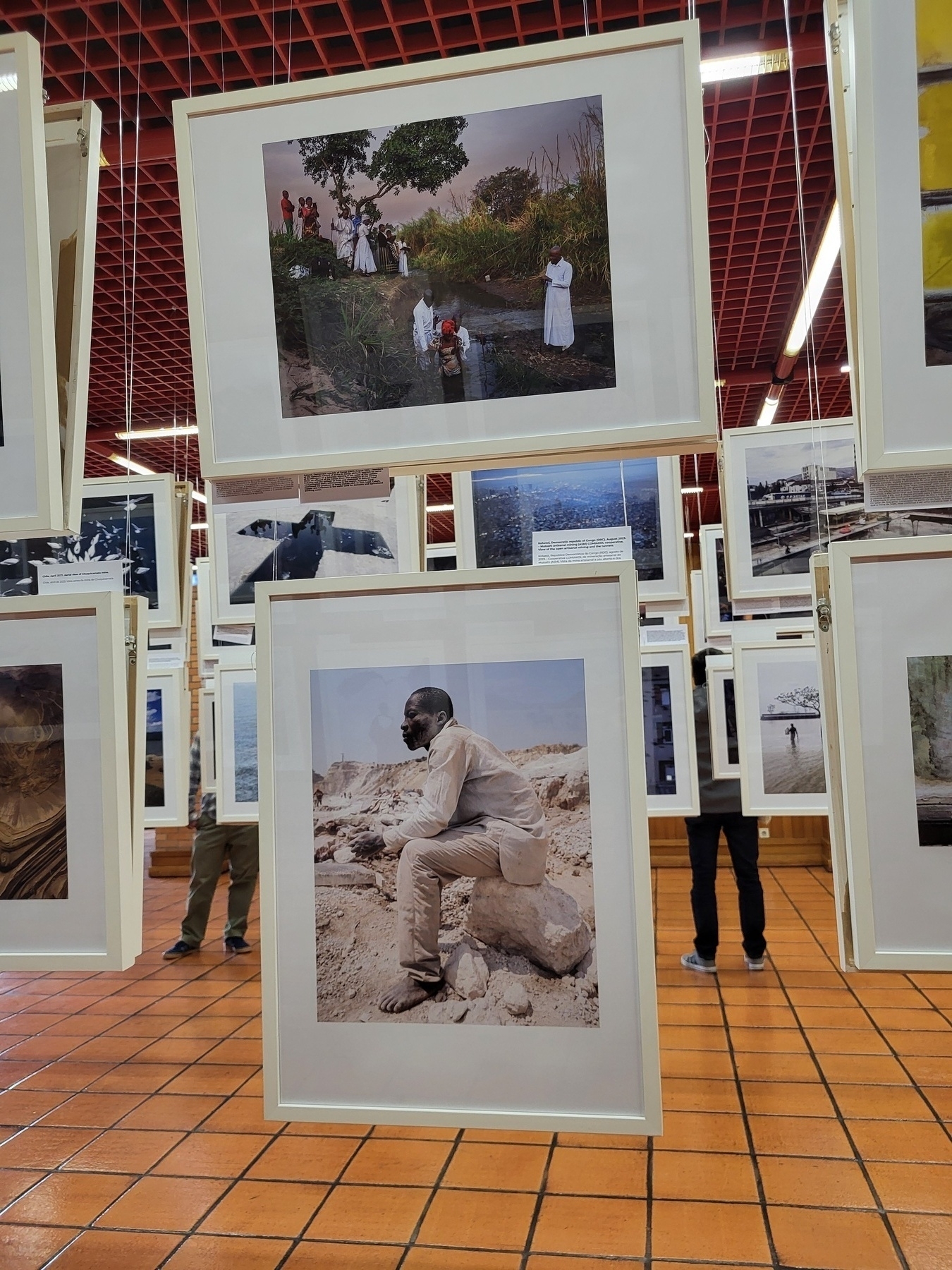 Framed photographs are suspended from the ceiling in a grid-like arrangement within an exhibition space.