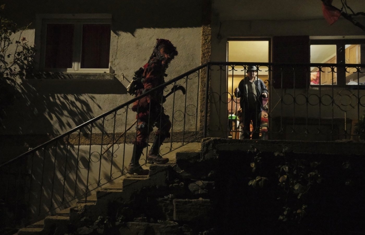 A person dressed in a dark, detailed costume with horns and holding a stick ascends stairs at night, while another person looks on from inside a lit room.