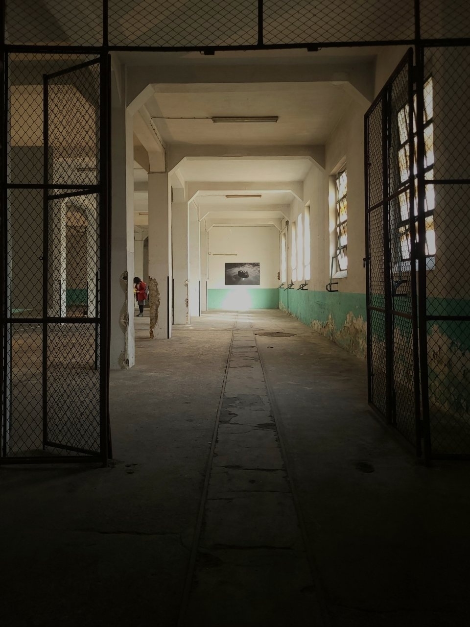 A long, industrial corridor with metal gates, peeling paint, and a lone person in the distance is illuminated by natural light streaming through barred windows.