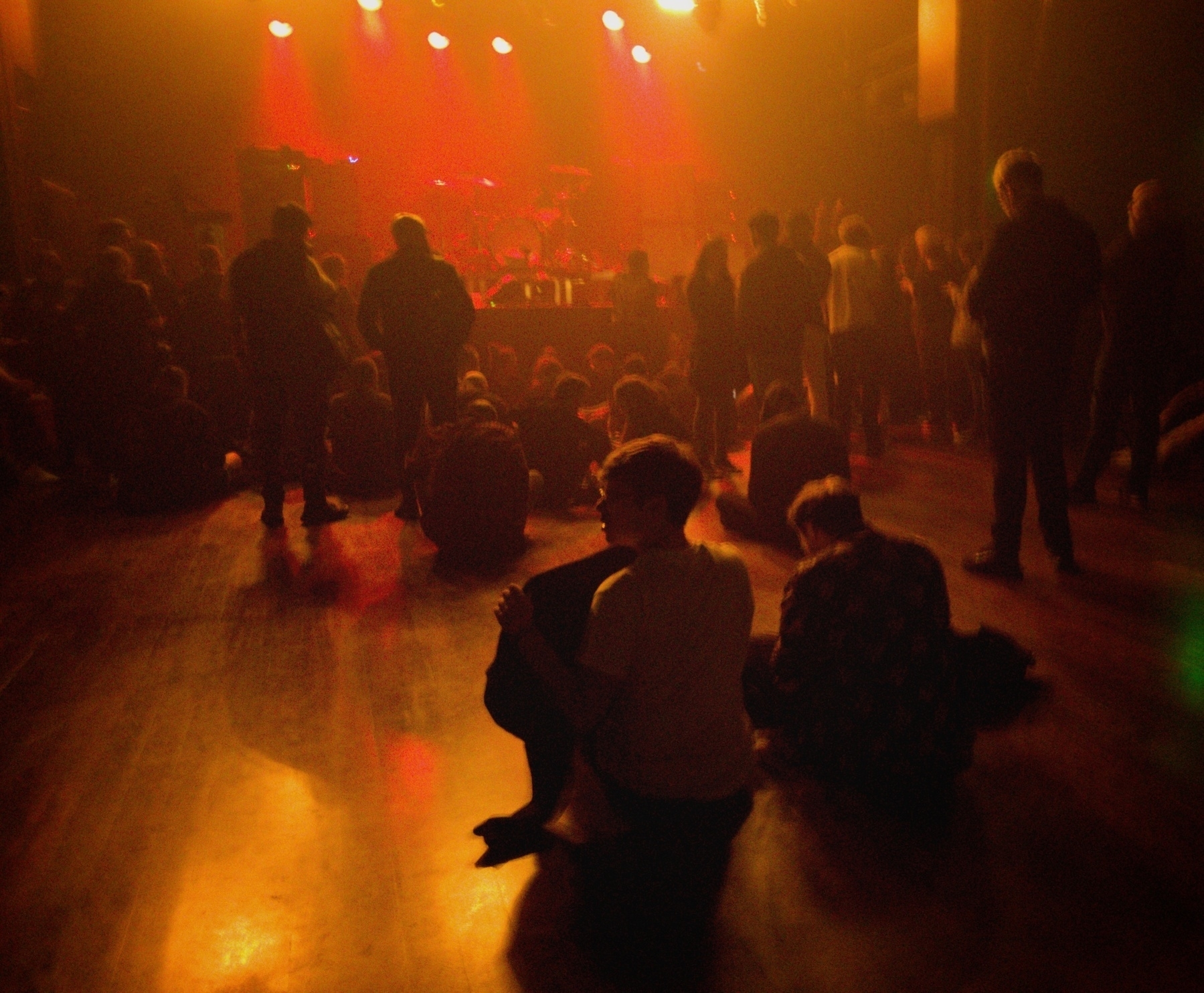 A group of people are gathered in a dimly lit concert venue, some sitting on the floor, facing a stage illuminated with orange lights.